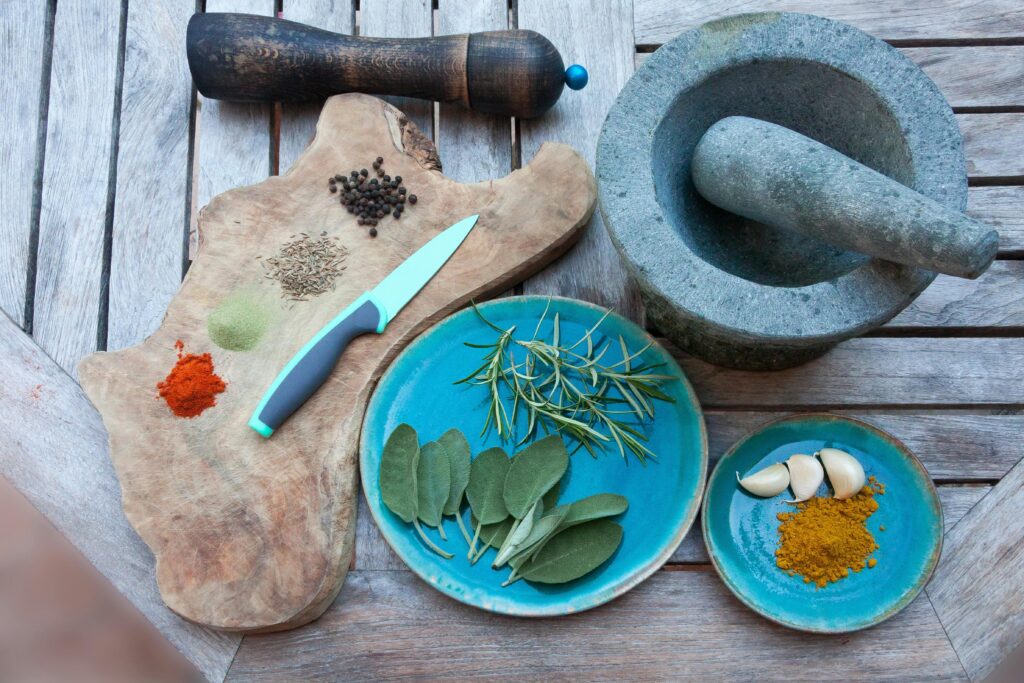 Herbs and spices are displayed on a cutting board and blue plates. They are surrounded by a grey mortar and pestle and wood pepper grinder.