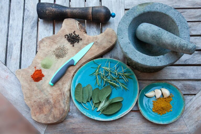 Herbs and spices are displayed on a cutting board and blue plates. They are surrounded by a grey mortar and pestle and wood pepper grinder.